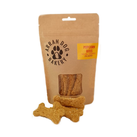 A brown paper bag labeled "Arran Dog Bakery" is open, revealing three bone-shaped Popcorn Bites Arran Dog Biscuits, natural vegetarian dog treats that are handmade in Brodick.