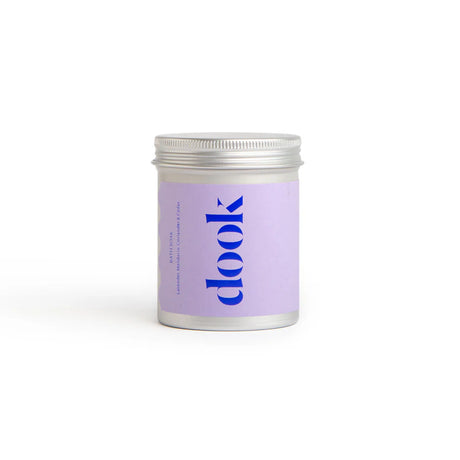 A small, cylindrical metal container with a screw-top lid and a lavender label boldly displaying "dook" in blue letters houses the "Lavender, Mandarin, Coriander & Cedar Bath Salt." Positioned against a plain white background, this soothing soak promises an indulgent escape.