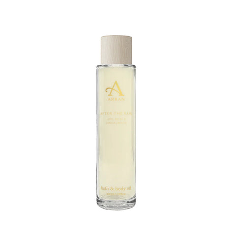 A 100ml clear glass bottle of Arran's "After The Rain" bath oil. The bottle features a light-colored, wooden-textured cap with gold lettering. The product is enriched with essential oils including lime, rose, and sandalwood. It is photographed against a white background.