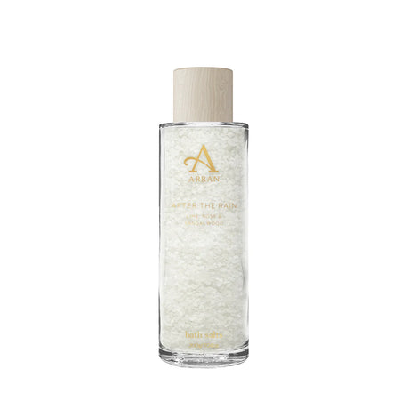 A tall, clear glass bottle filled with "After The Rain" Bath Salts (225g) features a wooden cap and gold lettering. The label highlights notes of lime, rose, and sandalwood.