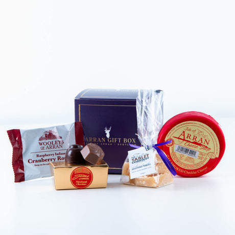 The Wee Favourites Arran Gift Box prominently features Isle of Arran delights: a red-wrapped Oak Smoked Cheddar, a package of cranberry rose, small containers of chocolates, and a bag of toffee, all elegantly arranged against a dark blue background.