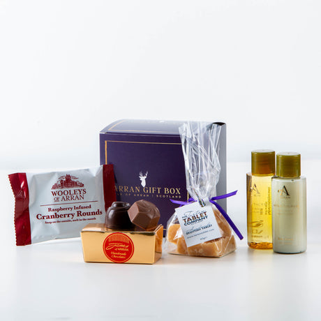 The Wee Dun Fionn Arran Gift Box provides a unique Isle of Arran-themed experience, including a packet of Woolley's cranberry rounds, gold-wrapped chocolates, fudge in clear packaging, and two small bottles of toiletries featuring the invigorating After The Rain fragrance.