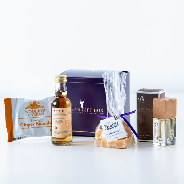 Wee Lochranza (10 Year Old Malt) Arran Gift Box, showcasing a small bottle of distinguished Arran Whisky, a package of Woolley's ginger rounds, a bag of Scottish tablet pieces adorned with a purple ribbon, and an elegant bottle of Lochranza Eau de Toilette, all elegantly arranged against a light background.