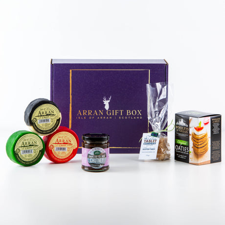 The Original Favourites Arran Gift Box, featuring a striking purple design, includes an assortment of foods: three Isle of Arran Cheddar cheeses (one herband one oak smoked, one mustard), a jar of caramelised onion chutney, a packet of oatcakes, and a bag of Scottish tablet.