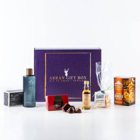 A selection of luxury items from The Fallen Rocks - Sannox Edition is displayed, including bath products, chocolates, a small bottle of Arran Whisky, ginger biscuits, and a packet of oatcakes, all set against a white background. The blue box features gold lettering.