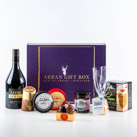 The Deluxe Arran Gold Lover Arran Gift Box features a bottle of Arran Gold Cream Liqueur, assorted cheeses, oatcakes, chutney, and chocolates, beautifully displayed in front of a striking purple box labeled "Arran Gift Box - Isle of Arran, Scotland.