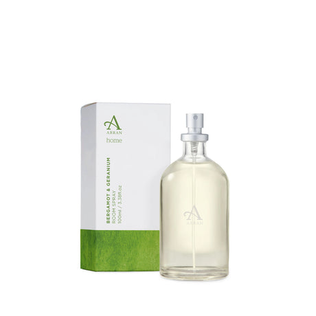 A transparent glass bottle of Bergamot and Geranium Room Spray 100ml, featuring a silver nozzle, is positioned beside its matching green and white box against a plain white background.