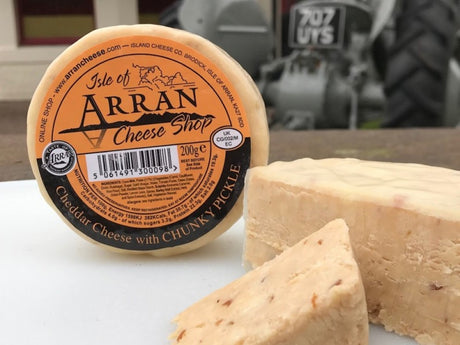 A round package of Chunky Pickle Cheddar from Isle of Arran is enticingly placed next to slices on a rustic cheeseboard, with a tractor blurred in the background.