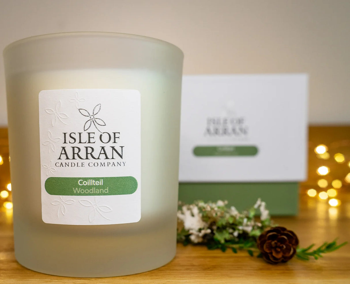 A frosted glass candle from the Isle of Arran Candle Company, labeled "Coilteill Woodland," is elegantly showcased in front of its packaging. The setting, rich with woody accents, features small decorations such as pinecones and greenery, enhanced by the soft glow of blurred fairy lights in the background.