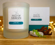 A frosted glass candle from the Isle of Arran Candle Company, named "Cuan-ard Stormy Seas," is positioned in front of its packaging on a wooden surface. The setting includes decorative elements like a stone, seaweed, and subtle string lights in the background, evoking the refreshing spirit of tumultuous seas.