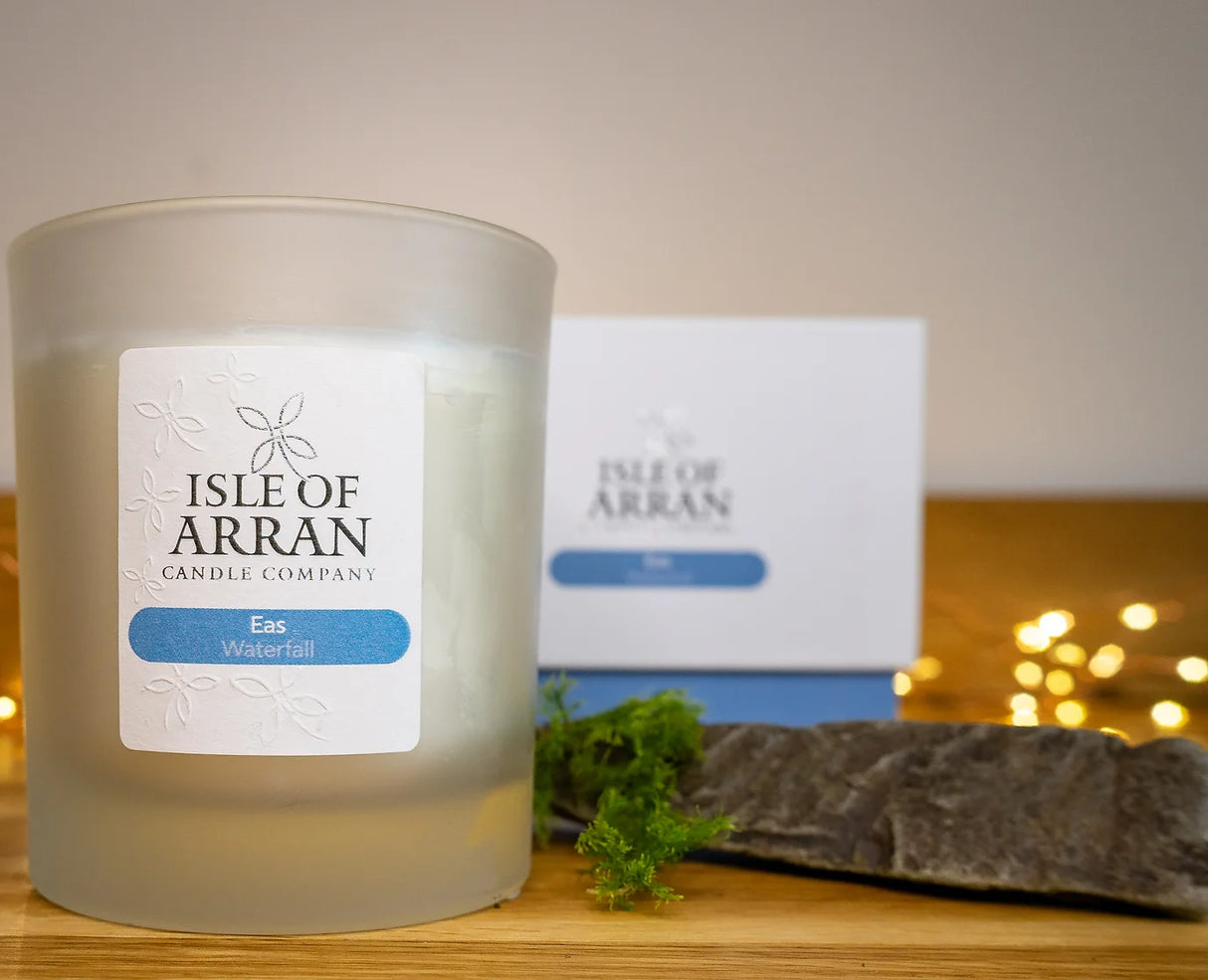 An Eas Waterfall Candle from the Isle of Arran Candle Company sits elegantly on a wooden surface. Its invigorating scent blends harmoniously with nature, accompanied by a nearby rock and greenery. A box in the background and gentle fairy lights enhance the cozy ambiance reminiscent of cascading waterfalls.