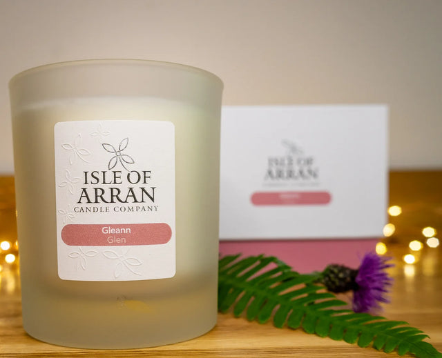 A frosted glass "Gleann Glen Candle" from the Isle of Arran Candle Company rests on a wooden surface. The scent emanating from it evokes vivid aromas, while a fern and purple thistle adorn the foreground. Fairy lights and the candle's branded box form a softly blurred background, reminiscent of Arran's stunning glens.