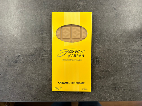 A yellow box labeled "James Chocolates Luxury Bar of Caramel Chocolate" displays visible caramel chocolate pieces through a clear window. The packaging indicates a weight of 100 grams, resembling an enticing caramel chocolate bar, elegantly placed on a dark surface.