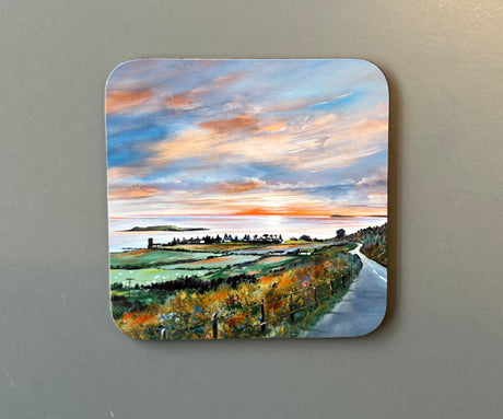 The CHARLES HERBERT COASTER by Charles Herbert showcases a scenic painting of a coastal landscape at sunset. The artwork depicts a winding road, lush greenery, a distant shoreline, and a sky graced with vibrant hues of orange, pink, and blue.