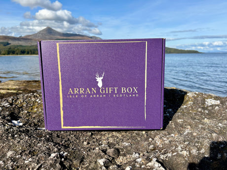 A box with a purple shade and golden text reading "Arran Gift Box - Medium Size Gift Box, Isle of Arran, Scotland" rests on rocky terrain near the water's edge, promising a delightful experience of Scottish delicacies. The backdrop features a serene lake, distant mountains, and a bright blue sky.