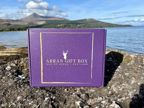 A large Arran Gift Box in purple, featuring "Isle of Arran, Scotland" in gold lettering, rests on a rock by a lakeside. Majestic mountains and a partly cloudy sky grace the background, creating an ideal setting for savoring exquisite Scottish delicacies.