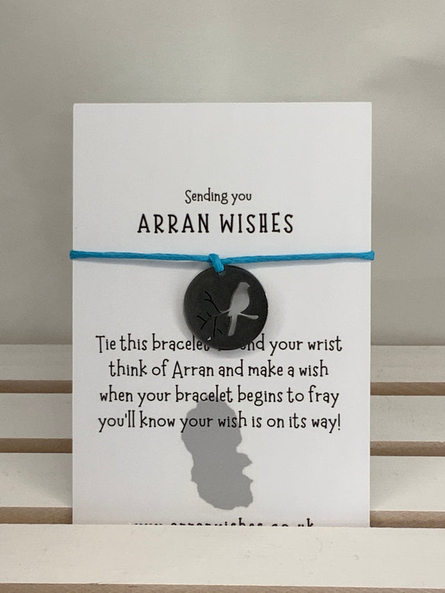 The "Arran Wish Bracelet - Shell" presents an Arran-inspired charm on a blue cord with a black circular emblem adorned with a bird. It encourages you to tie the bracelet, make a wish, and anticipate its magic as it starts to fray.