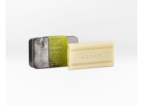 A bar of Machrie Men's Soap 200g is showcased in front of its rectangular metallic green and silver packaging. The light beige, triple-milled soap has the word "Arran" elegantly embossed on it, highlighting its quality craftsmanship.