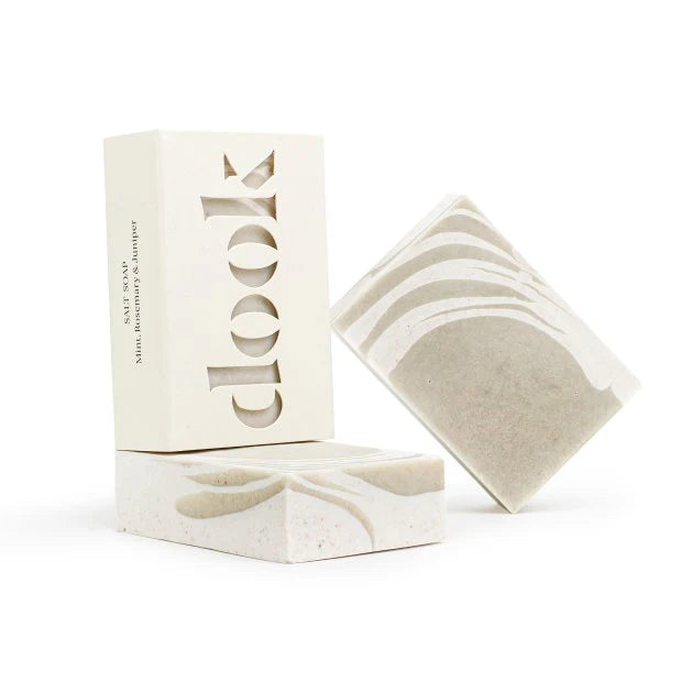 Two sleek, rectangular bars of Mint, Rosemary & Juniper Salt Soap with a minimalist design rest on a white surface. The soap showcases marbled swirls in neutral tones. One bar is partially wrapped in white packaging with cut-out design spelling "clook.