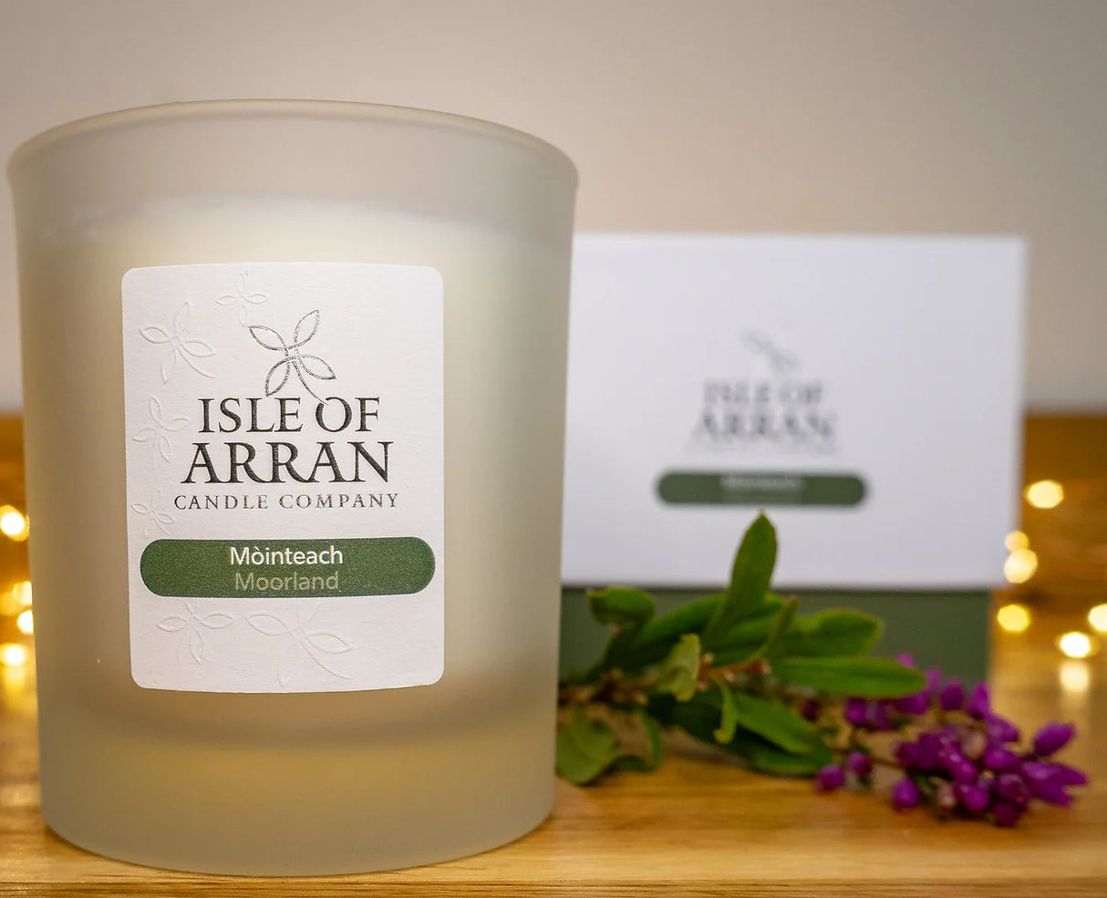 A frosted glass "Mòinteach Moorland Candle" from the Isle of Arran Candle Company releases herbaceous aromas, accompanied by a matching box in the background. The scene is enhanced by purple flowers and greenery on the wooden surface, with warm fairy lights evoking serene moorlands and adding to a cozy atmosphere.