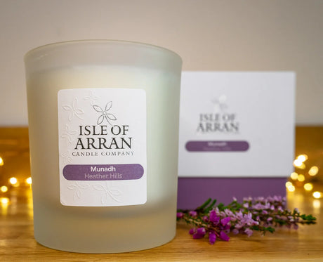 A frosted glass "Munadh Heather Hills Candle" from the Isle of Arran Candle Company radiates a floral aroma. It is gracefully positioned in front of a coordinating box, surrounded by delicate purple flowers and gentle fairy lights that capture the enchanting beauty of heather-covered hills.