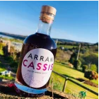 A 20cl bottle of Arran Cassis, brimming with the richness of blackcurrants, is placed on a wooden surface outdoors. The label displays "Arran Cassis." Behind it, a picturesque green landscape extends under a clear blue sky, making it ideal for preparing an exquisite Kir Royale.