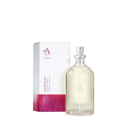 A clear glass bottle of Ultimate Fig Room Spray 100ml, featuring a silver spray nozzle, is positioned beside its matching box. The box is predominantly white with a pink bottom section and displays the brand’s logo.