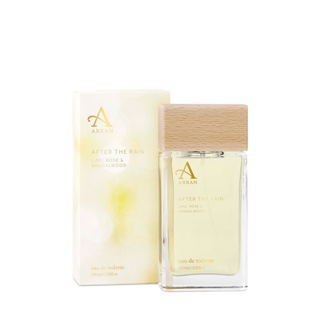 A bottle of After The Rain Eau De Toilette 100ml from Arran Sense of Scotland sits beside its box. The minimalist designs on both the box and bottle are complemented by a wooden cap. The label reveals a fragrance blend of lime, rose, and sandalwood.