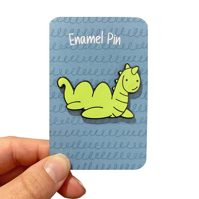 A card is held, displaying a "Nessie Enamel Pin Badge" sculpted as an adorable smiling cartoon dinosaur in green. The card's blue backdrop showcases squiggly lines with "Enamel Pin" at the top, and a rubber clutch secures the pin well.