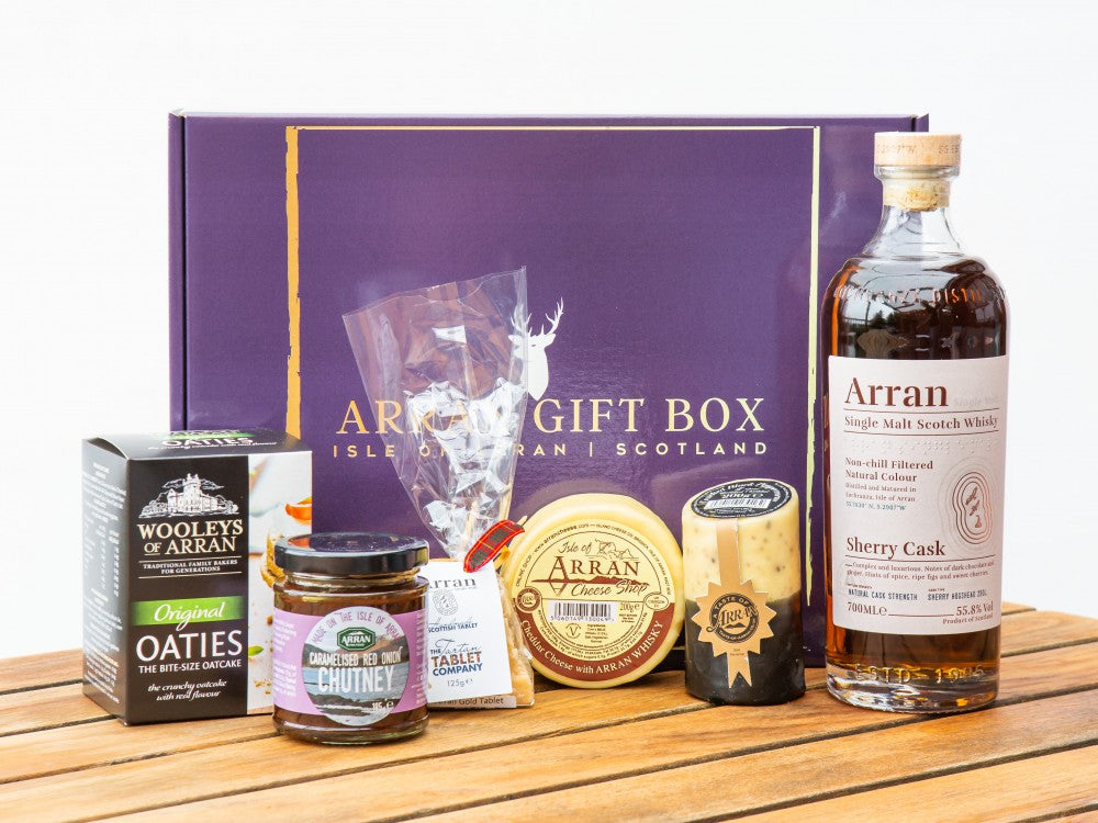 A purple gift box titled "Deluxe Whisky Lover (Sherry Cask Finish Malt) Arran Gift Box" is filled with Scottish treasures: a bottle of Sherry Cask Finish Single Malt, oatcakes, chutney, two cheeses, and sweets, all elegantly displayed on a wooden surface.