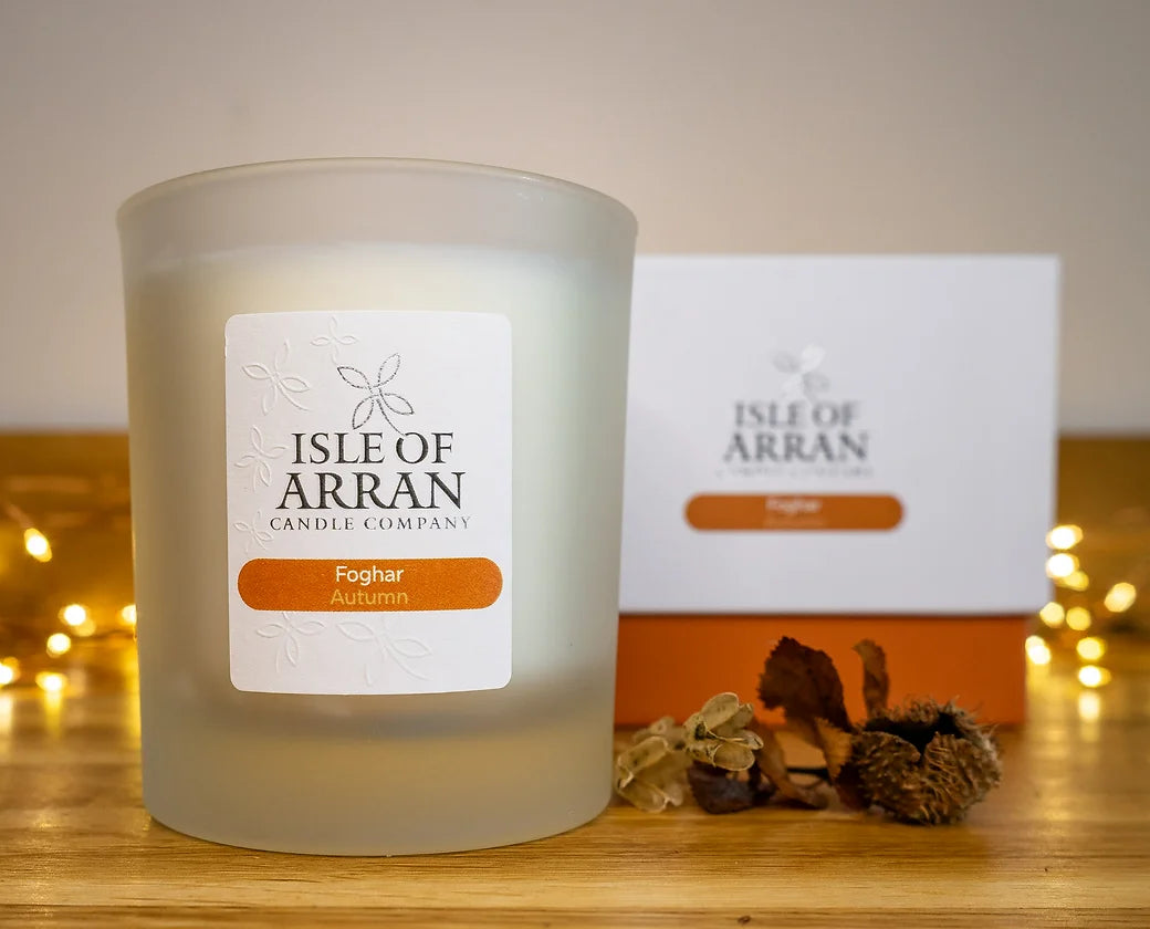 In the foreground, a frosted glass Autumn Candle labeled "Foghar - Autumn Candle" captures attention. Handcrafted in Scotland by Isle of Arran Candle Company, it is accompanied by its matching box and surrounded by small autumnal decorations and string lights on a wooden surface, creating an enchanting and cozy ambiance.