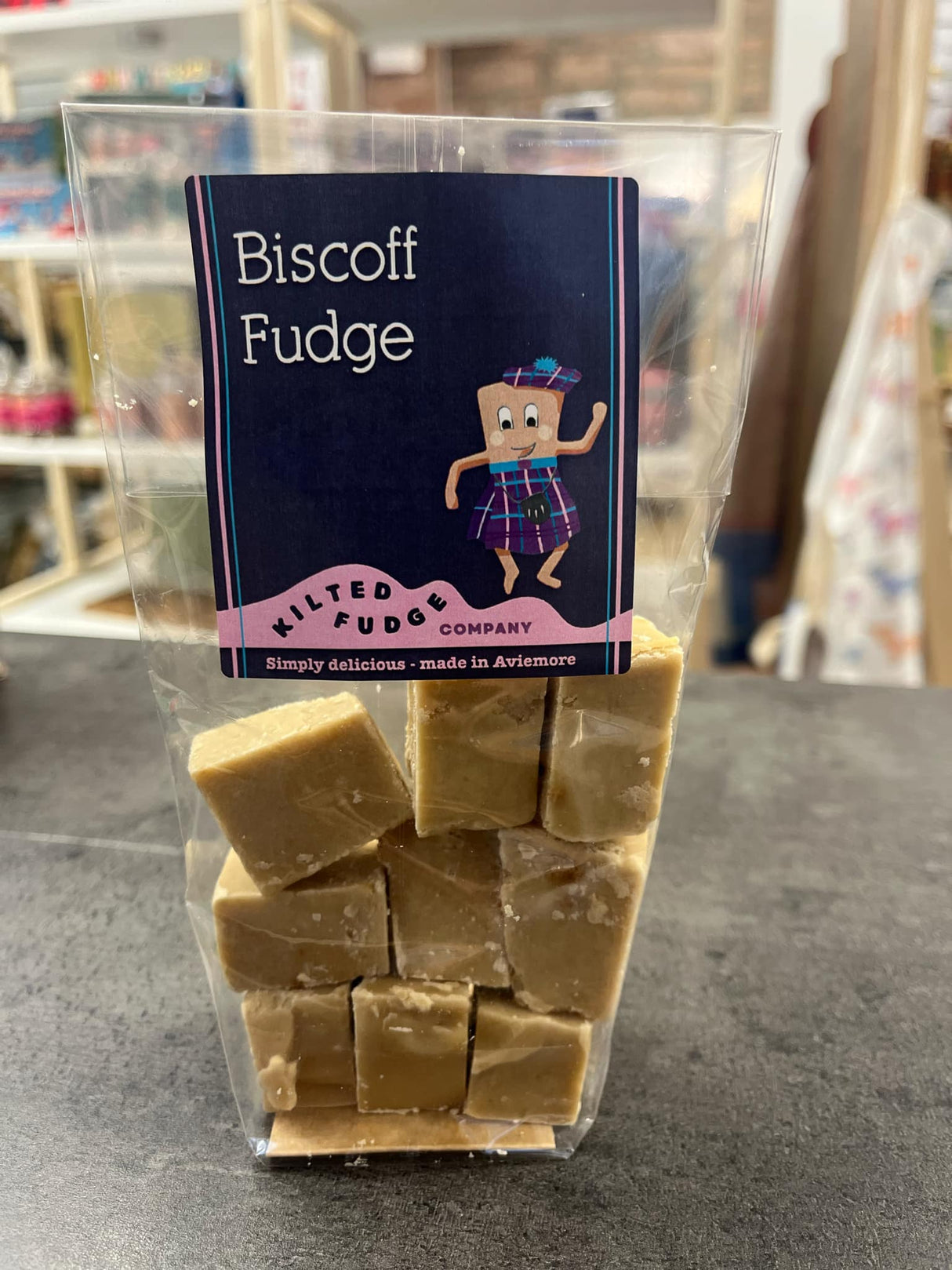 A clear bag containing Kilted Fudge Company's Biscoff Fudge is showcased on a countertop, offering enticing notes of Biscoff spread. The packaging showcases a cartoon fudge character confidently wearing a kilt.