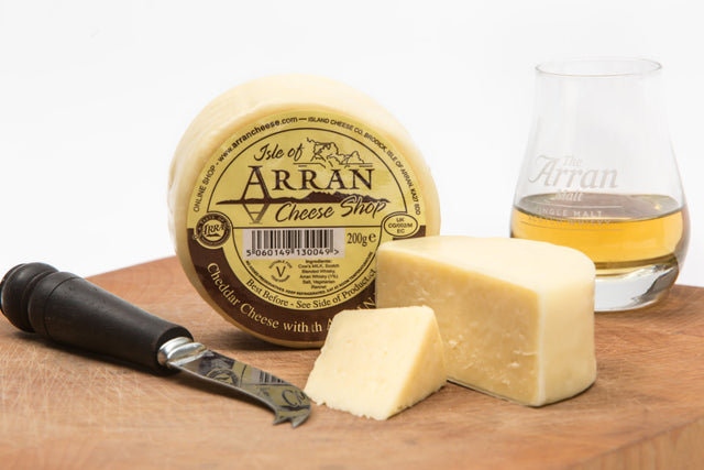 A round block of Arran Whisky Cheddar rests on a wooden board, enhanced by a wedge cut out. Next to it lies a cheese knife and a glass of Arran Malt, embodying the spirit of the Scottish Highlands. The label prominently displays the branding of the Isle of Arran Cheese Shop.