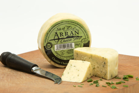 A round block of Chive Flavoured Cheddar cheese from Isle of Arran rests on a wooden board with a slice cut out. Nearby, a cheese knife sits surrounded by chopped herbs and several Arran Oatcakes. The cheese label is prominently displayed, showcasing its name and details.