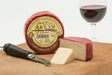 A block of Claret Cheddar is partially sliced next to a cheese knife, resting on a cheeseboard. The cheddar is encased in red wax, with a glass of red wine elegantly posed in the background on a wooden surface.