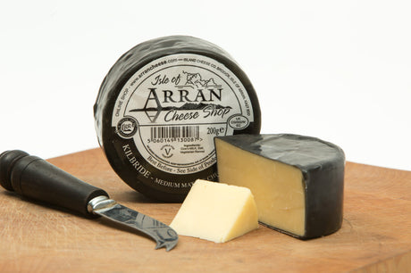 A round block of Kilbride (Plain) Cheddar cheese, creamy and matured, partially sliced, sits on a wooden board. A cheese knife rests beside it. The Cheddar is wrapped in black wax, and the label on the front is visible.