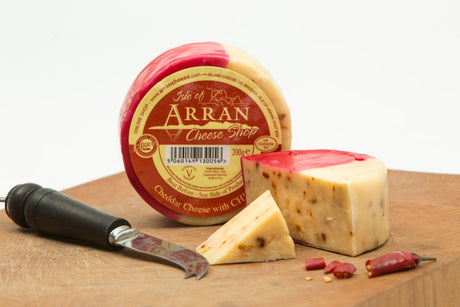 A round block of Chilli Flavoured Cheddar cheese from Scotland sits on a wooden board, partially unwrapped to reveal sliced pieces speckled with bright chilli seeds. Nearby, a small cheese knife and a few chili peppers enhance the rustic presentation.