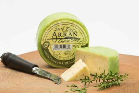 A round block of Herb Flavoured cheddar, encased in a green wax coating, sits elegantly on a wooden board. A wedge is cut out, revealing its creamy interior enriched with the finest herbs. Nearby, a cheese knife and sprigs of thyme complete this delightful display of Scottish cheddar elegance.