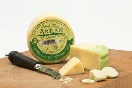 A wheel of Smokey Garlic Cheese from the Isle of Arran Cheese Shop is partially sliced to reveal garlic bits, displayed on a wooden cheeseboard alongside a cheese knife and garlic cloves. The cheese boasts a distinctive green wax coating.