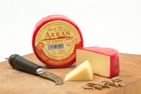 A wheel of Oak Smoked Cheddar, encased in red wax, is artfully arranged on a wooden board. It is accompanied by a cheese knife and wedge, with small pieces of shaved oak wood providing a rustic accent to the creamy cheddar display.