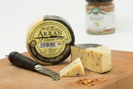 A wheel of "Arran Mustard flavoured Cheddar" with a slice that reveals wholegrain mustard seeds sits enticingly on a wooden board. Complementing this flavored cheese, a black-handled knife rests nearby, and in the background, a jar of Arran Mustard enhances the gourmet allure.