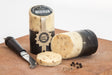 Two wheels of Cracked Black Peppercorn Cheddar, coated in black wax, are showcased on a wooden board. One wheel is sliced open, revealing its peppercorn-speckled interior. Accompanying the cheese are a cheese knife and scattered coriander seeds.