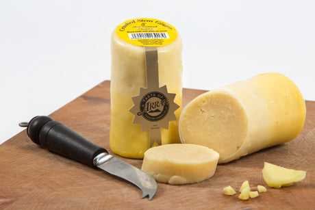 A block of Crushed Stem Ginger Cheddar, coated with wax, is sliced open on a wooden board. A cheese knife with a black handle sits nearby. A round piece of cheese is in the foreground, and the alluring label proudly displays its heritage on the wax.